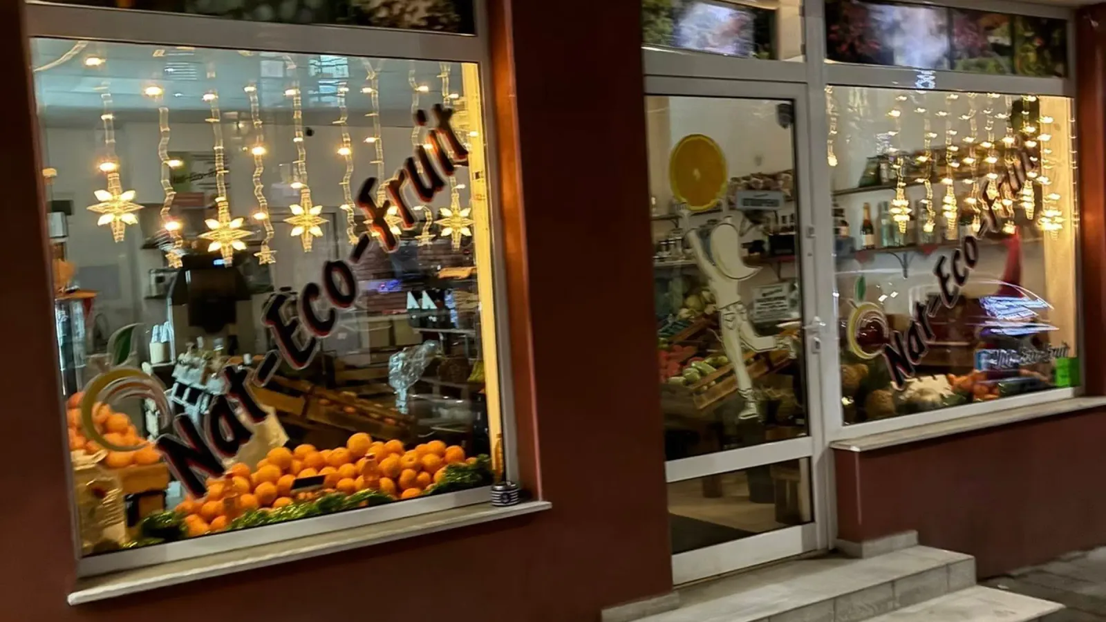 Фасадата на Nat-Eco-Fruit вечерта, с осветени витрини, букви на бранда и кошница с цитруси